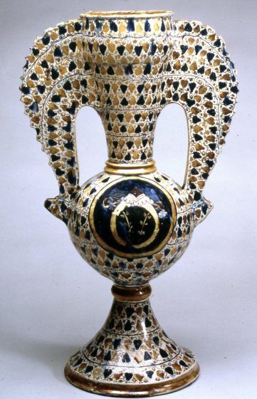 A white vase with multicolored geometric patterns painted on. A coat of arms is featured in the center of the vase. Art like this is a part of the legacy of ceramics from Islamic Spain.