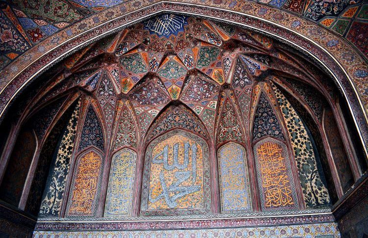 A wall of a mosque, showing verses of the Quran in Arabic as well as multicolored paintings of plants.
